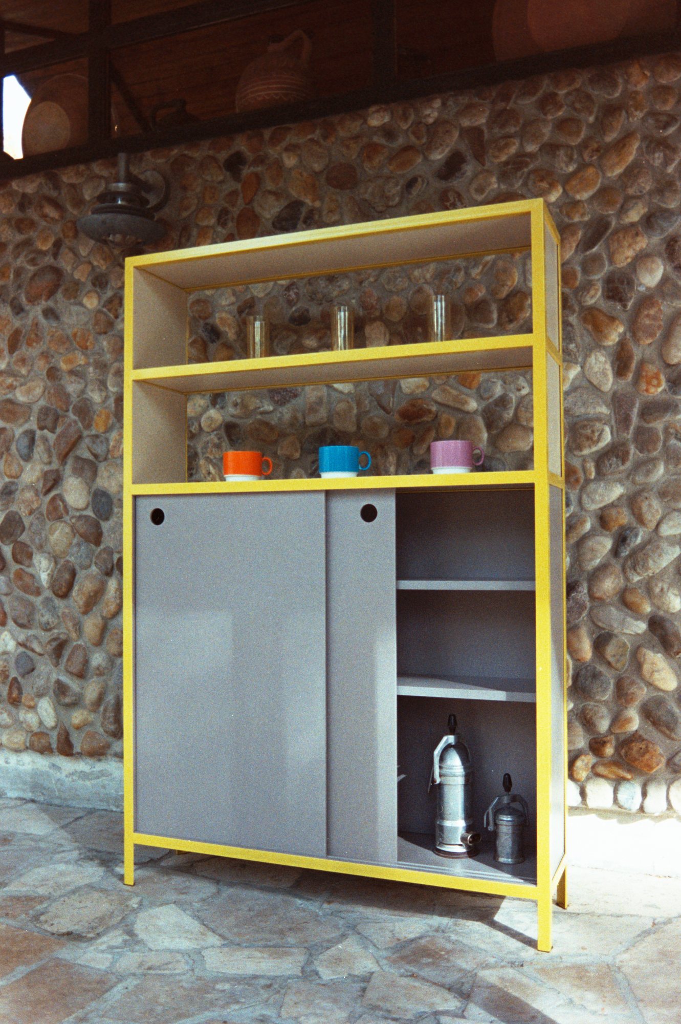 Frame Shelf with a yellow steel frame and grey sliding doors against a stone wall